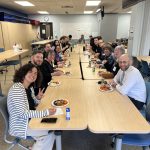 A group photo in which people are seated on both sides of a very long table in a large, brightly-lit classroom