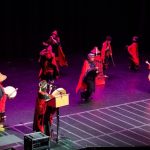 A photo of the Haida dance group G’aahlandee Xander dancing on a stage with purple lighting, red and black clothing, and a dark backdrop